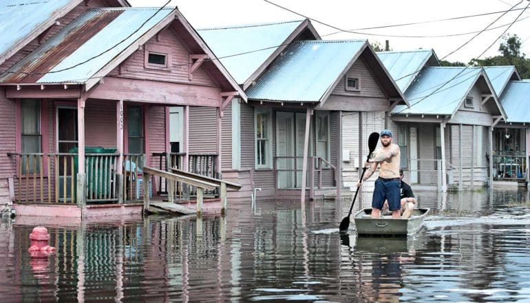 A man in a row boat rows down a flooded street past houses with water coming up to the front porch