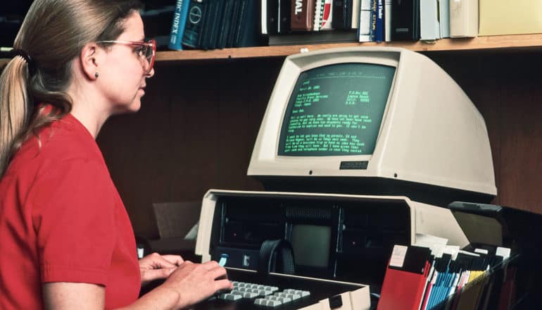 woman in "Sally Jesse" glasses uses 1980s computer in office