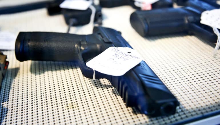A black handgun sits on display in a gun store with a white price tag on it and other guns in the background