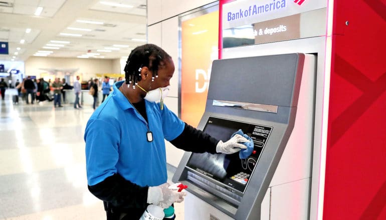 A man in a blue shirt, gloves, and a face mask wipes down an ATM machine at the airport