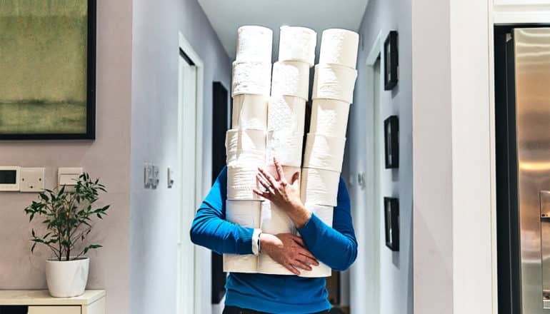 A person carries almost 2 dozen toilet paper rolls in a large stack down their hallway