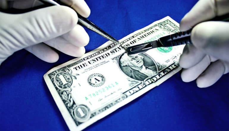 A surgeon with white gloves uses a scalpel to slice a dollar bill in half as it sits on a blue surface