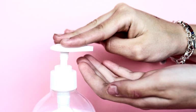 A pair of hands press a hand soap pump down against a pink background