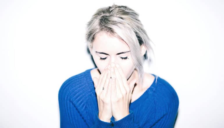A young woman in a blue sweater covers her face while sneezing or coughing
