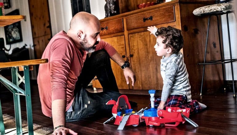 A father speaks with his son, with both sitting on the kitchen floor as the son plays with toys