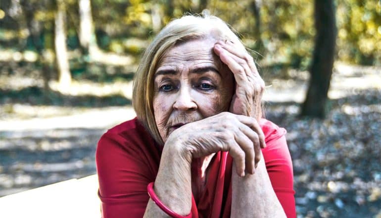 An older woman touches her temple and leans on her hand while sitting on a park bench outside