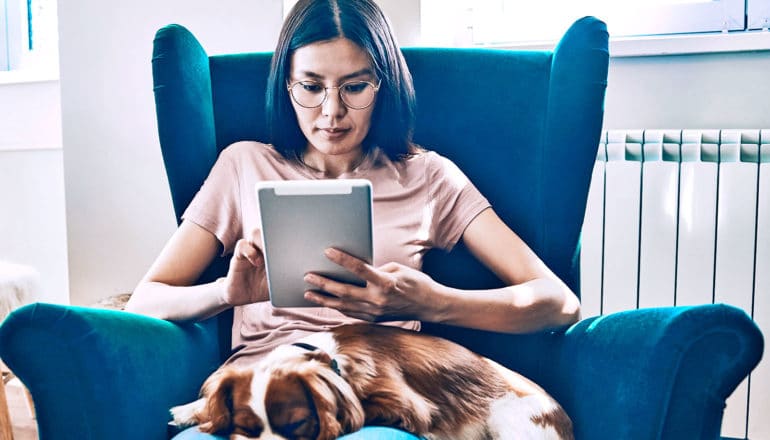 A woman sits in a blue chair and reads a tablet computer, while her dog sits on her lap