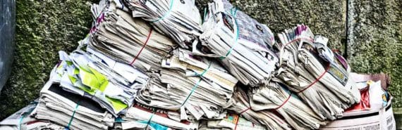 Stacks of newspapers lean to the right, slumping onto a box filled with more newspapers