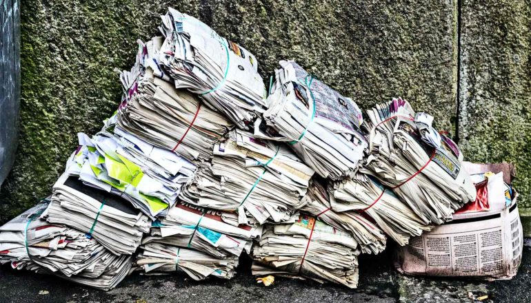 Stacks of newspapers lean to the right, slumping onto a box filled with more newspapers