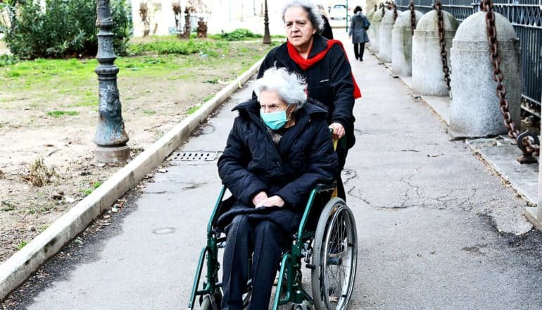 An older woman pushes another wearing a medical mask down a small side street in a wheelchair