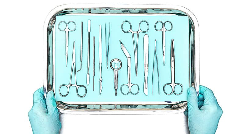 Gloved hands hold a tray of surgical instruments against a white background