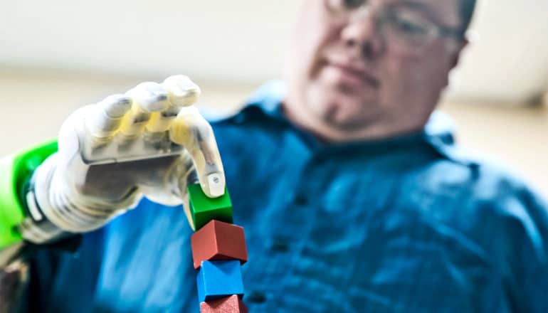 Hamilton uses the white, slightly translucent prosthetic hand to pick up a small green block sitting on a tower of other blocks between thumb and index finger