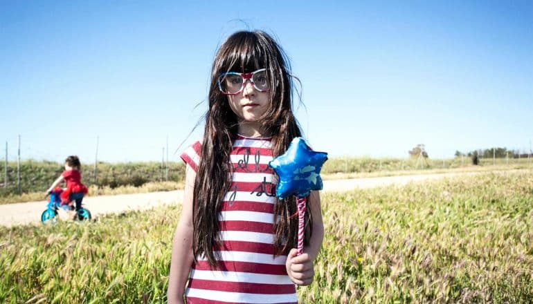 A young girl in a red and white striped t-shirt and glasses stands in front of a field and dirt road while holding a magic wand toy with a blue star at its end.