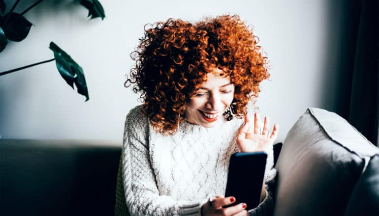 A woman with red hair smiles and waves into her phone camera while using Facetime