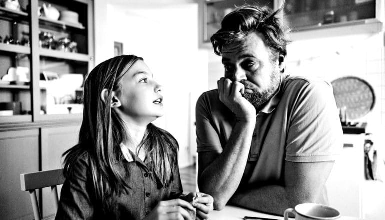 A father and daughter look at each other while sitting at the kitchen table. She looks hesitant, while he rests his face on his hand