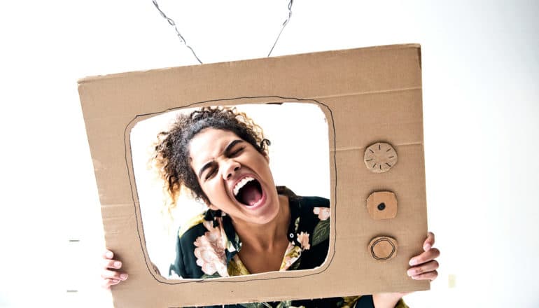 A woman holds up a cardboard TV around her face and screams through what would be the screen