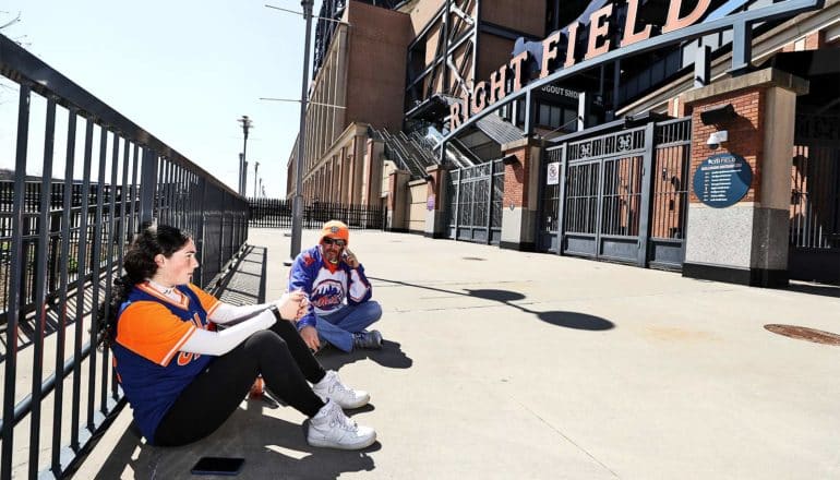 A father and daughter wearing New York Mets gear sit outside Citi Field looking sad and bored