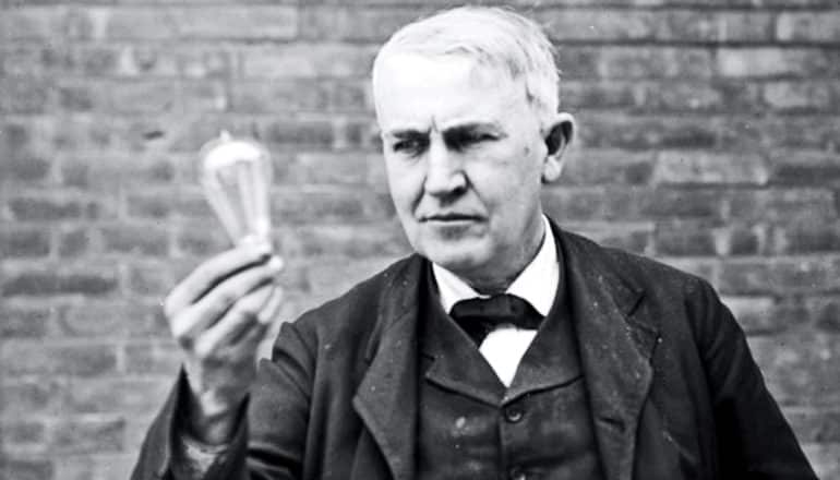 Thomas Edison holds a light bulb as he stands against a brick wall