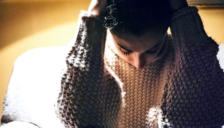 A young woman puts her hands on the back of her head as she looks down while sitting on the couch