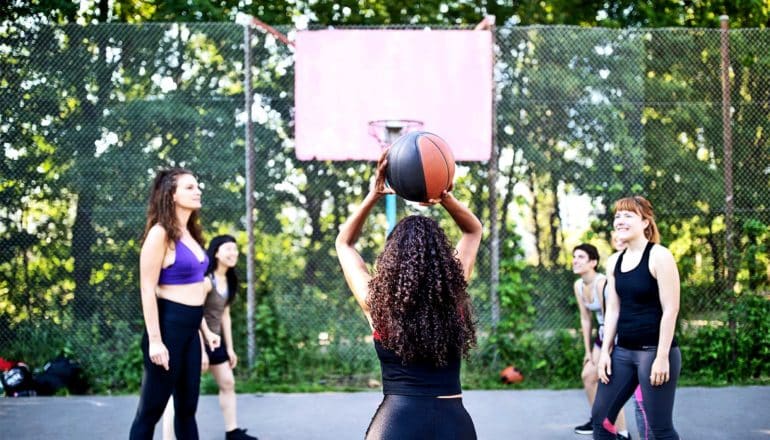 A woman is about to shoot a free throw. Other players stand on the sides of the key. The backboard of the hoop is pink and there are trees in the background behind it