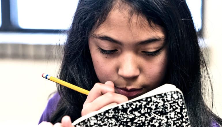 A young girl holds a pencil to her face while looking down at her black-and-white composition book