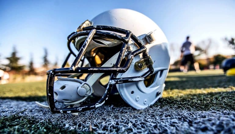 A white football helmet sits on the grass of a football field, with players, trees, and blue sky in the background