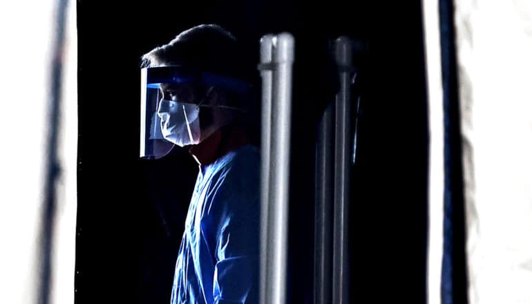 A health care worker stands in a dark tent wearing a face mask and blue protective gown