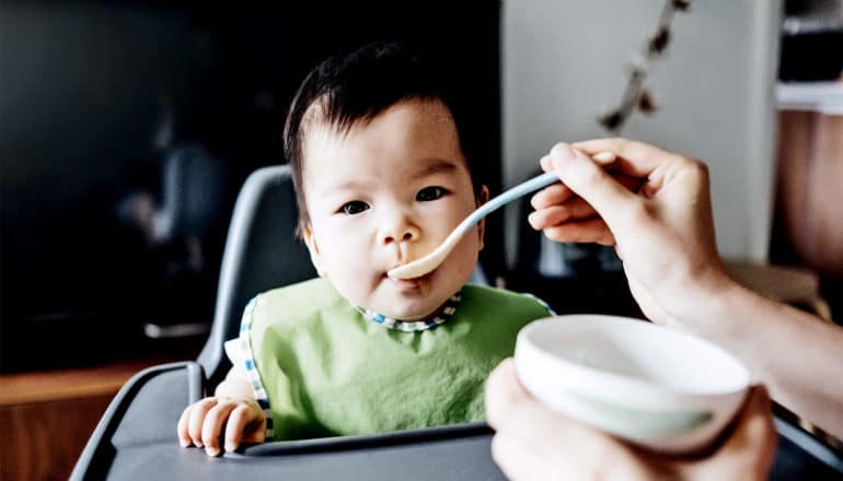 A parent spoons food into her babies mouth. The baby has a green bib on and a spoon in their mouth.