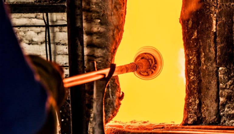 A glass blower holds a long rod with a blob of molten glass on the end of it, which is sitting in a bright orange furnace
