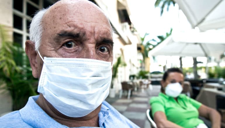 An older bald man wears a white medical mask, with a concerned look on his face. A woman in a medical mask sits at the same table behind him