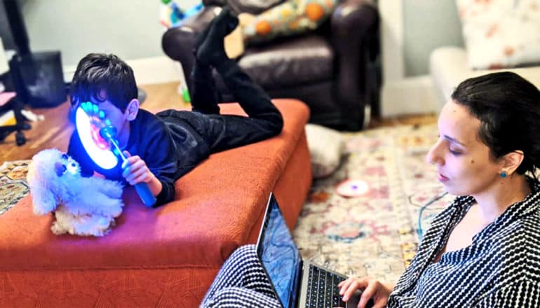 A child lays on an an ottoman examining a stuffed animal while his mother sits on the ground and looks at her laptop