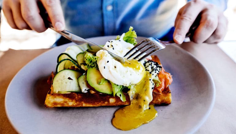 A man eats a breakfast plate that includes a waffle, egg, and avocado