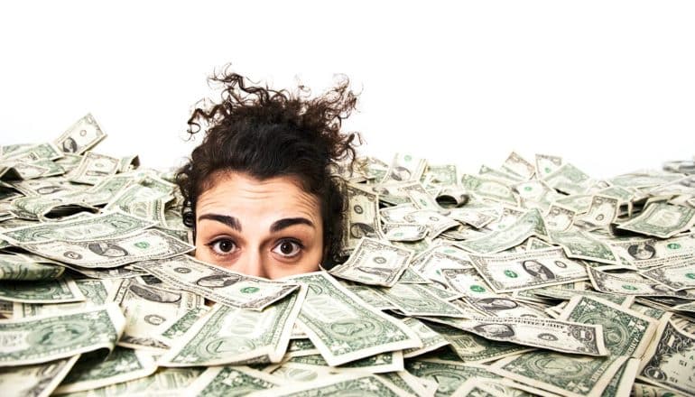 A young woman peeks out of a sea of dollar bills with an overwhelmed or stressed look