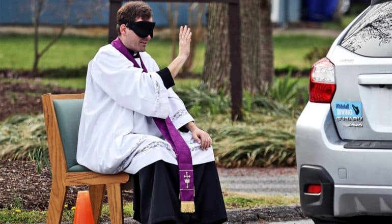 A blindfolded priest in white robes with a purple sash holds his hand up to make the sign of the cross towards a car near him in the parking lot