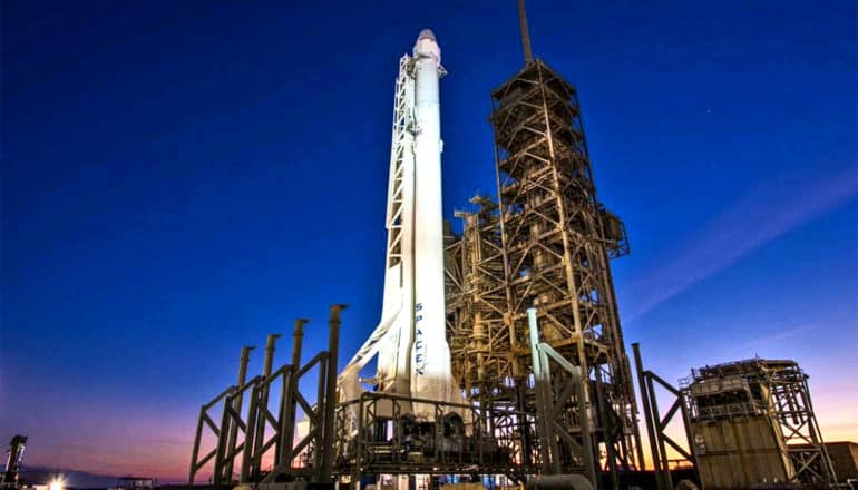 A SpaceX rocket stands ready for lift-off against a deep blue sky at dusk