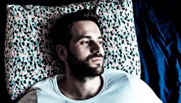 A young bearded man in a white t-shirt lays in bed wide awake