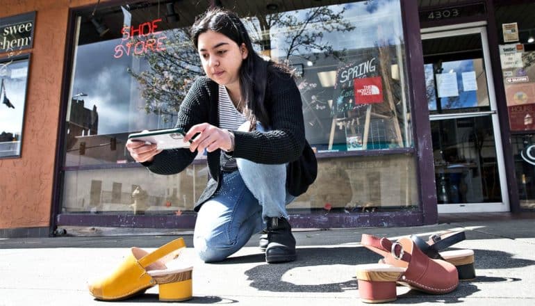 A young woman kneels outside a shoe store to take photos of shoes on the sidewalk