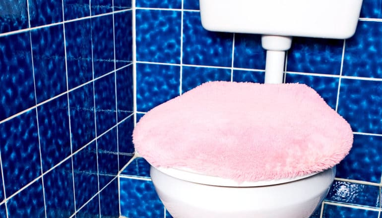 A white toilet with a fuzzy, pink lid cover sits in front of blue tiles