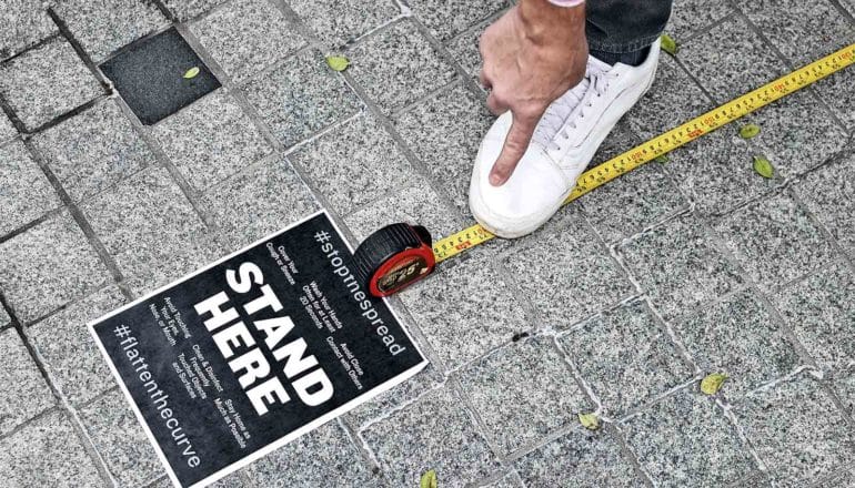 A person measures 6 feet with a tape measure, steps on the yellow tape with a white shoe, and points at the spot. A black sign reads "Stand Here."