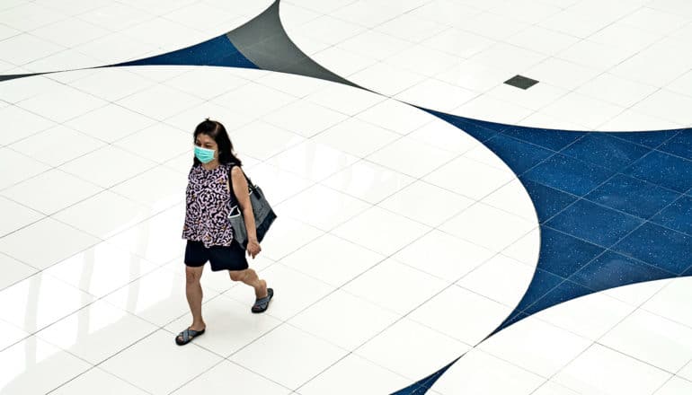 A woman walks over a white and blue tiled floor while wearing a face mask and carrying a purse