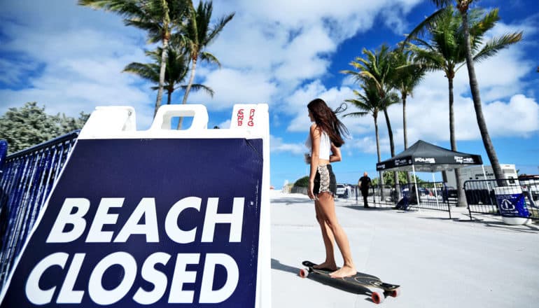 A young woman rides a longboard down a beach-side sidewalk past a sign in the foreground that says "Beach Closed." A police tent stands near the beach under palm trees and blue sky