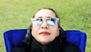 A woman sits in a blue chair outside while wearing sunglasses and looking up at palm trees and blue sky
