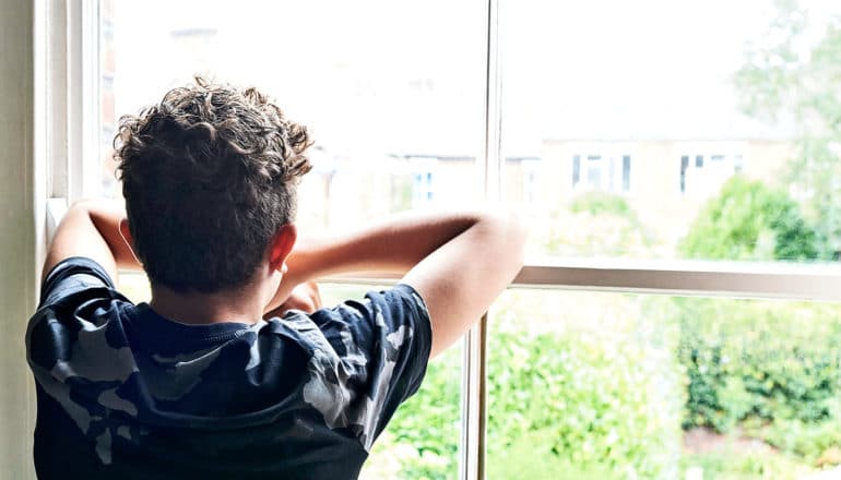 A young teen in a camo t-shirt looks out a closed window