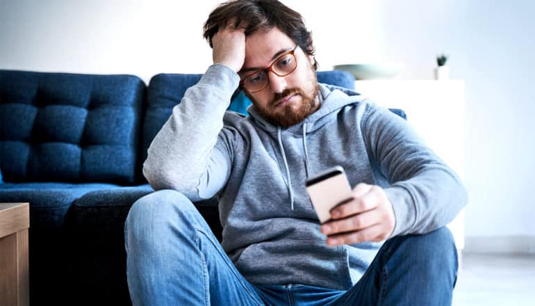 A man looks worried as he reads his phone while sitting on the floor in front of a blue couch