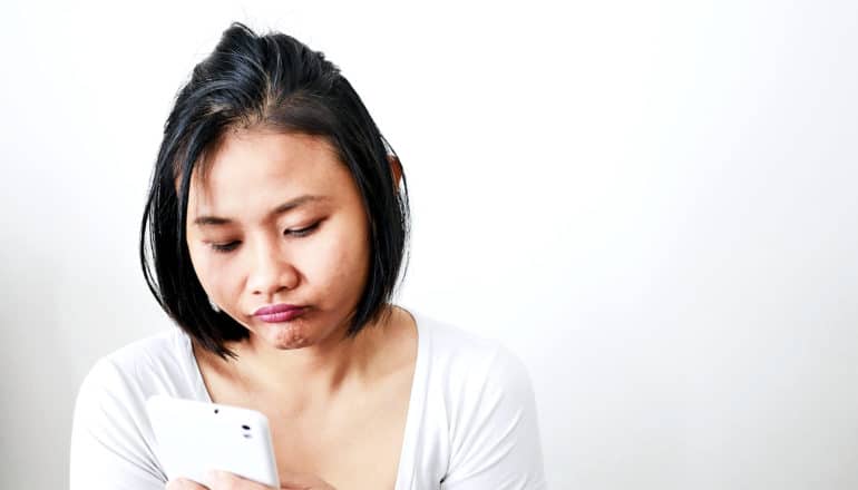 A young woman in a white shirt makes a frustrated and bored face while looking at her phone