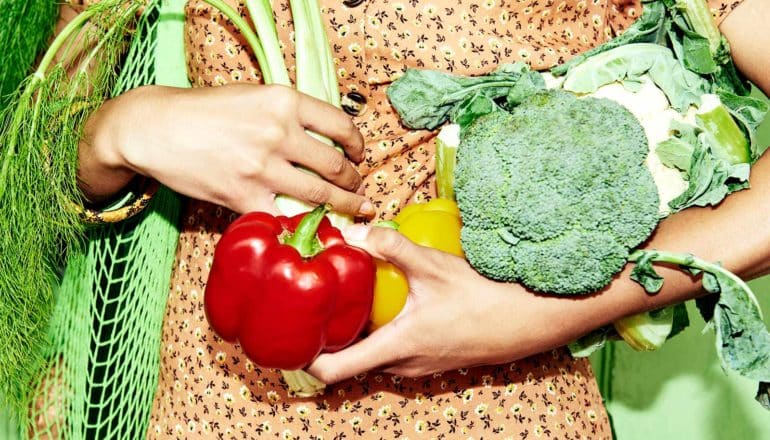 A woman holds different kinds of vegetables, including a red bell pepper, broccoli, and cauliflower, along with a green grocery bag made of netting