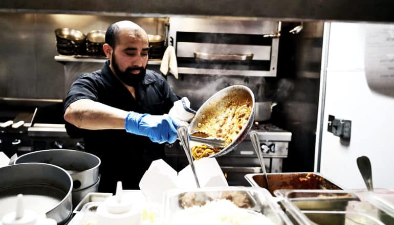 A man cooks in a restaurant kitchen wearing blue gloves. He's scraping food from a pan into a container