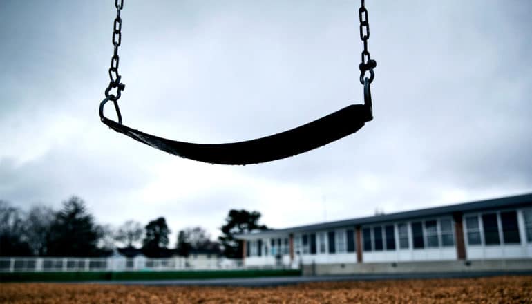 A swing hangs at an empty school yard with gray clouds in the background