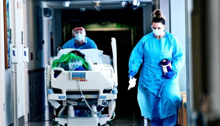 Health care workers in blue protective equipment push a COVID-19 patient down a hallway on a gurney