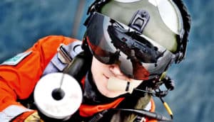 A man in search-and-rescue gear ascends a rope wearing a helmet and visor, in which you can see the helicopter above him reflected